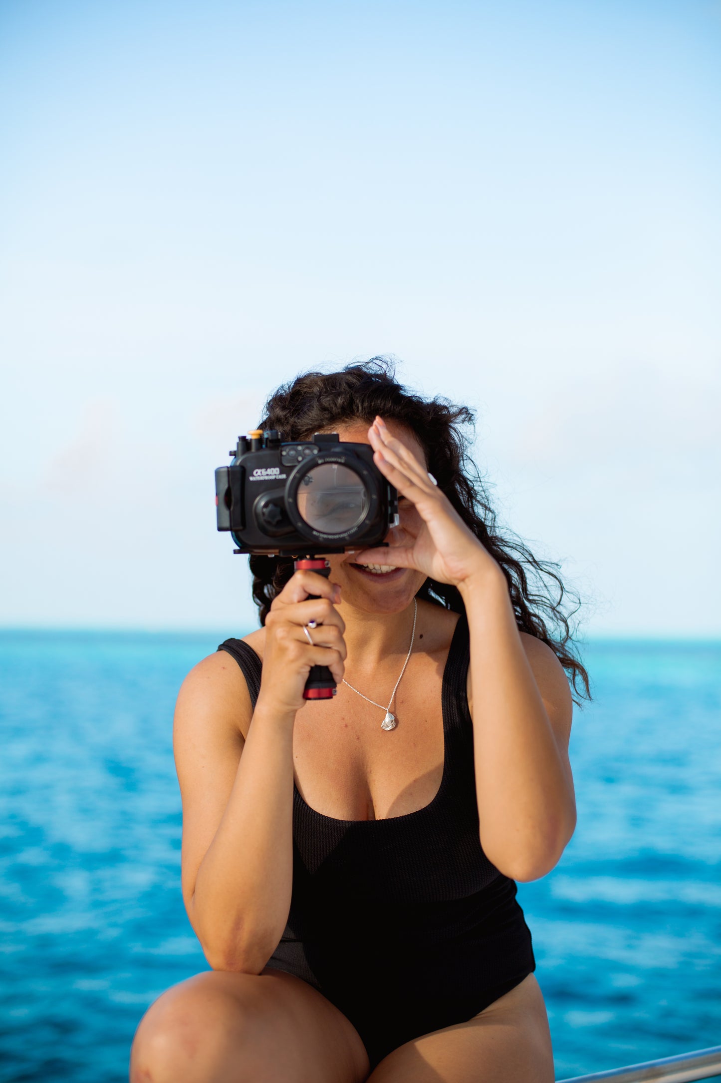 Model wears large silver oyster lid necklace on boat with ocean behind her and camera in hand
