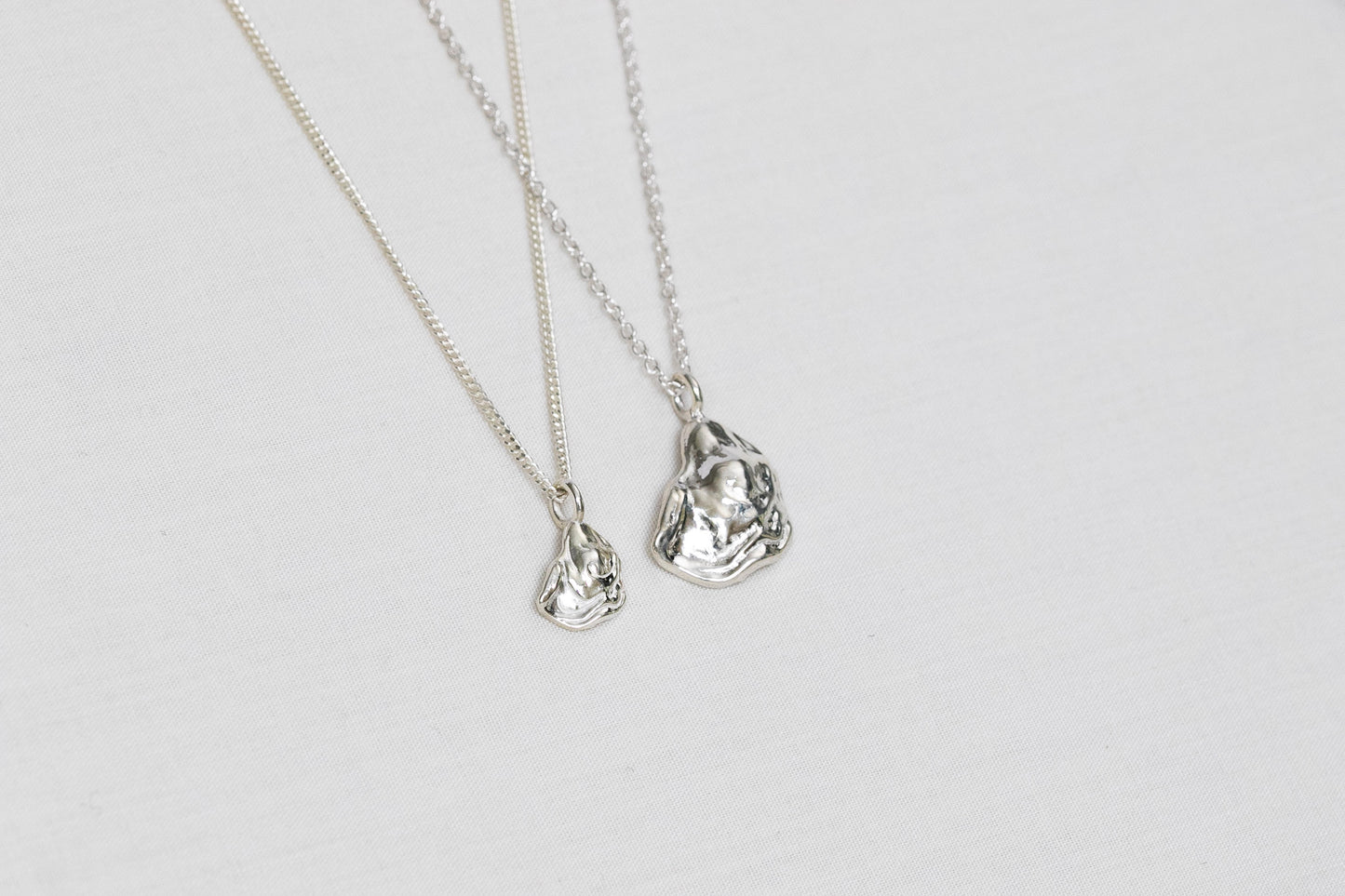 Two oyster necklaces lay side by side, one mini size and one larger size, both handmade from sterling silver