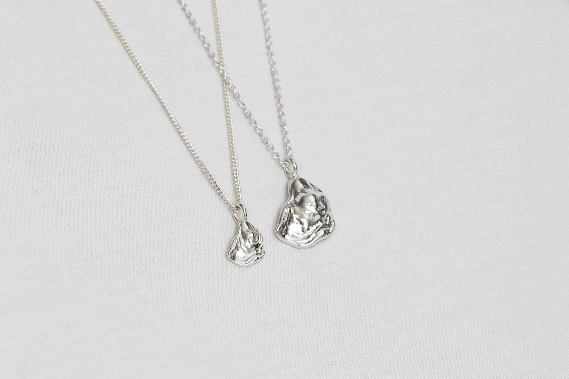 Two oyster necklaces lay side by side, one mini size and one larger size, both handmade from sterling silver